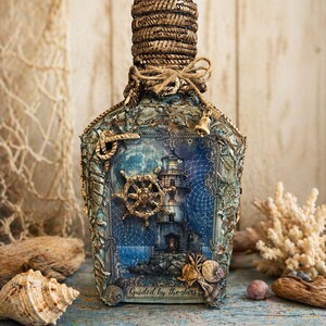 Puede incluir: Botella decorativa con temática náutica. La botella presenta una escena de faro, un timón de barco y está adornada con cuerda, conchas y el texto "Guided by the stars". El tapón de la botella está envuelto en cuerda.
