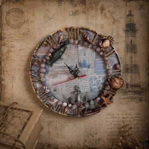 May include: A round wall clock featuring a collage of blue and white nautical-themed paper, Roman numerals, and a border decorated with small seashells, a green fish, a crab, a starfish, a small boat, and a silver anchor at the bottom.