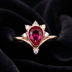 May include: A rose gold ring with a teardrop-shaped, deep red gemstone at its centre. The gemstone is encircled by smaller, clear accent stones, in a crown-like arrangement. The ring is set against a black backdrop.