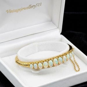 Może przedstawiać: Złota bransoletka typu bangle z zawiasem, ozdobiona rzędem jedenastu owalnych opali w szlifie kaboszonowym. Bransoletka posiada delikatny złoty łańcuszek zabezpieczający i jest umieszczona w białym pudełku z nadrukiem Vintagejewellery511 wewnątrz.