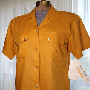Puede incluir: Una camisa de lino de manga corta de color mostaza con botones dorados y dos bolsillos de solapa. La camisa tiene cuello y está en un maniquí. La etiqueta dice "SK & COMPANY, Made in the USA."