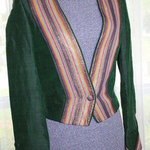 Op de afbeelding: Een groene fluwelen blazer met een kleurrijke gestreepte bies. De bies heeft een verscheidenheid aan kleuren, waaronder goud, paars, roze, oranje en blauw. De blazer heeft een enkele knoopsluiting.