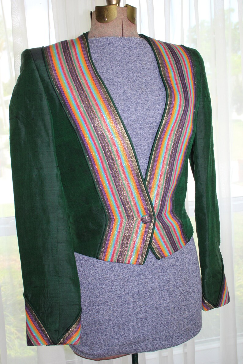 Op de afbeelding: Een groene fluwelen blazer met een kleurrijke gestreepte bies. De bies heeft een verscheidenheid aan kleuren, waaronder goud, paars, roze, oranje en blauw. De blazer heeft een enkele knoopsluiting.