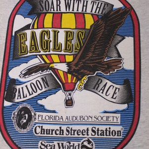 May include: A gray t-shirt with a colorful logo featuring a hot air balloon, an eagle, and the text "Soar with the Eagles Balloon Race". The logo also includes the text "Florida Audubon Society", "Church Street Station", and "Sea World Orlando, Florida".