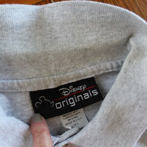 May include: Gray polo shirt with a black tag that says "Disney Originals" and has a Mickey Mouse head logo. The tag also includes care instructions and size information: "Machine wash, Tumble dry warm, RN 51926, Made in USA, X-L."