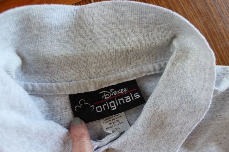 May include: Gray polo shirt with a black tag that says "Disney Originals" and has a Mickey Mouse head logo. The tag also includes care instructions and size information: "Machine wash, Tumble dry warm, RN 51926, Made in USA, X-L."