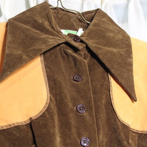 May include: A vintage brown corduroy jacket with a large collar and tan suede-like panels. The jacket has three buttons down the front and is hanging on a metal hanger. The stitching is visible around the tan panels.