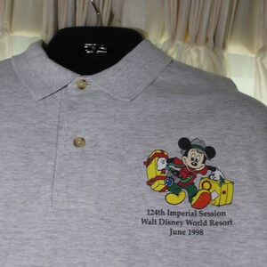 May include: Gray polo shirt with a cartoon image of Mickey Mouse carrying luggage. The text on the shirt reads "124th Imperial Session Walt Disney World Resort June 1998".