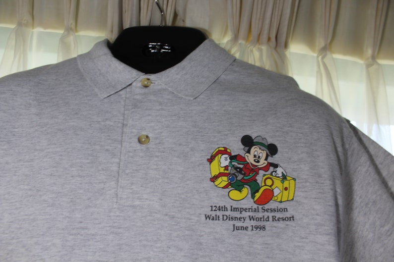 May include: Gray polo shirt with a cartoon image of Mickey Mouse carrying luggage. The text on the shirt reads "124th Imperial Session Walt Disney World Resort June 1998".