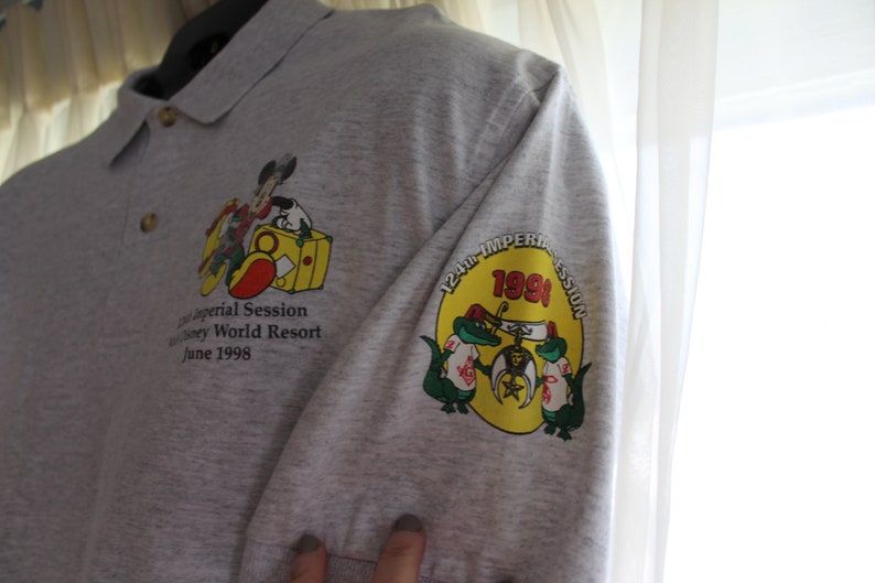 May include: Gray polo shirt with a yellow, red, and green embroidered patch on the right sleeve. The patch features the text "124th Imperial Session 1998" and two alligators. The left side of the shirt features a smaller embroidered patch with Mickey Mouse and the text "124th Imperial Session Disney World Resort June 1998."