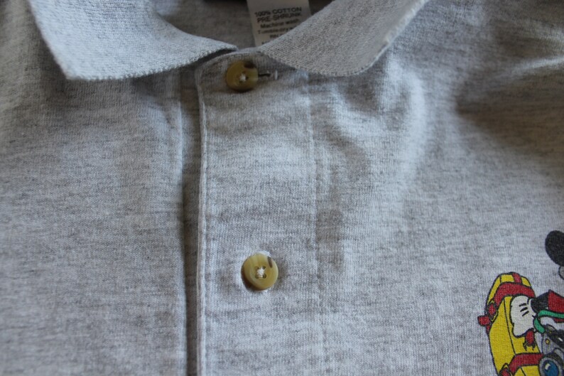 May include: A gray polo shirt with a button-up closure and a small Mickey Mouse design on the right side. The shirt is made of 100% cotton.
