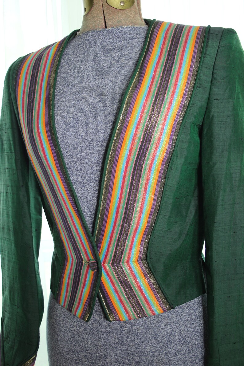 Op de afbeelding: Een groene blazer met een kleurrijke gestreepte bies. De bies is gemaakt van meerdere kleuren, waaronder goud, paars, oranje en blauw. De blazer heeft een enkele knoopsluiting.