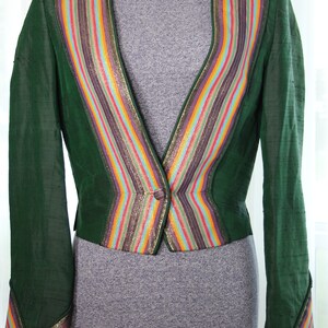 Op de afbeelding: Een groene blazer met een kleurrijke gestreepte bies. De bies heeft gouden, rode, oranje, gele, blauwe, groene en paarse strepen. De blazer heeft een enkele knoopsluiting.