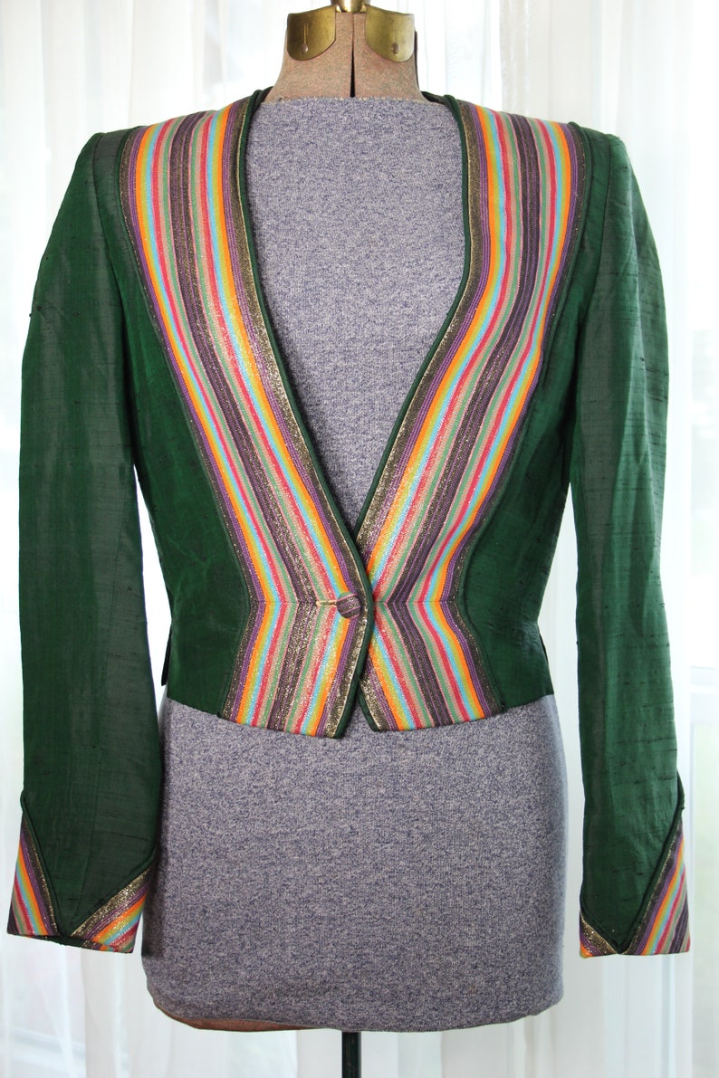Op de afbeelding: Een groene blazer met een kleurrijke gestreepte bies. De bies heeft gouden, rode, oranje, gele, blauwe, groene en paarse strepen. De blazer heeft een enkele knoopsluiting.