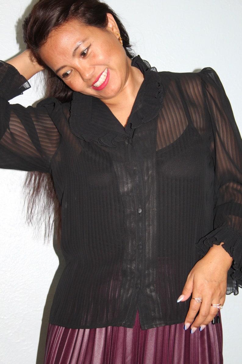 May include: A black sheer blouse with a ruffled collar and long sleeves. The blouse has a button-up front and is paired with a burgundy pleated skirt.
