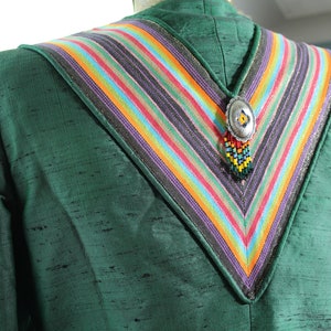 Op de afbeelding: Een groene blazer met een kleurrijke gestreepte kraag en een zilveren hanger met kralen die eraan hangen.