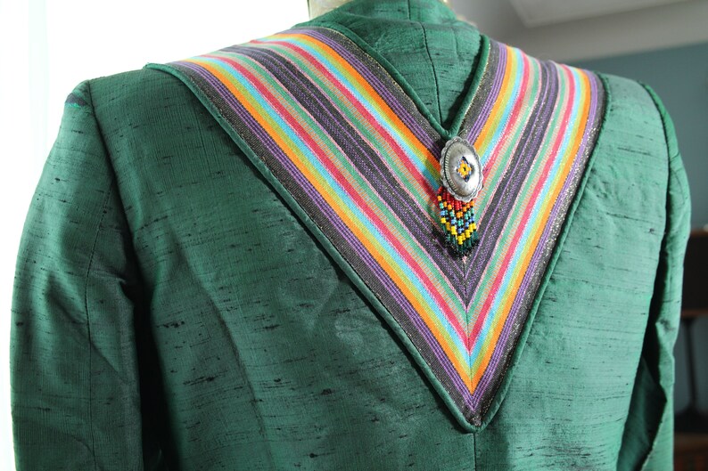 Op de afbeelding: Een groene blazer met een kleurrijke gestreepte kraag en een zilveren hanger met kralen die eraan hangen.