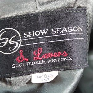 Op de afbeelding: Een zwart label met witte stiksels en de tekst "Show Season" en "d. Lavers Scottsdale, Arizona". Het label heeft ook een witte cirkel met de letters "GS" erin. Een kleiner wit label met de tekst "Dry Clean Only" is zichtbaar onder het grotere label.