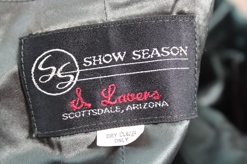 Op de afbeelding: Een zwart label met witte stiksels en de tekst "Show Season" en "d. Lavers Scottsdale, Arizona". Het label heeft ook een witte cirkel met de letters "GS" erin. Een kleiner wit label met de tekst "Dry Clean Only" is zichtbaar onder het grotere label.