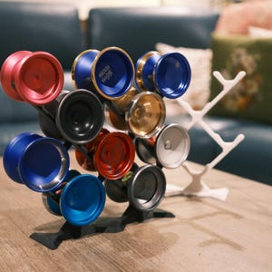 Puede incluir: Un expositor con una colección de yo-yos coloridos. Los yo-yos son de varios colores, incluyendo rojo, azul, negro y dorado. El expositor es blanco y negro, y los yo-yos están dispuestos de manera atractiva.