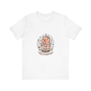 Puede incluir: Camiseta blanca con una ilustración de dibujos animados de un animal marrón bebiendo té boba. El boba está en una taza transparente con una pajita rosa. El texto en la camiseta dice "Boba Time" y "Sip Happens."