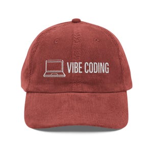Peut inclure: Casquette de baseball en velours côtelé rouge brique avec le texte blanc "VIBE CODING" et un dessin d'ordinateur portable blanc. La casquette a une visière incurvée et un dessus arrondi.