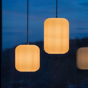 Op de afbeelding: Drie hanglampen met een warm, gloeiend licht. De lampen hebben een geribbeld, cilindrisch ontwerp en hangen aan zwarte snoeren tegen een donkerblauwe achtergrond. De lampen hebben een zachte, gebroken witte kleur.