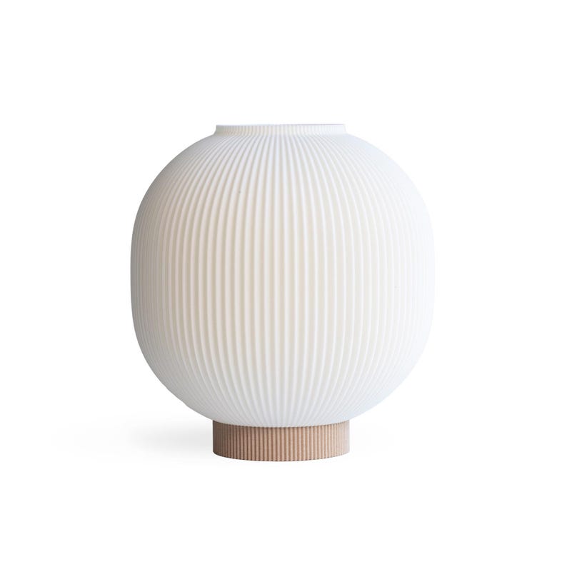 May include: A white, ribbed ceramic vase with an oval shape. The vase sits on a light brown, cylindrical base. The vertical ribs create a textured surface. The design is modern and minimalist.