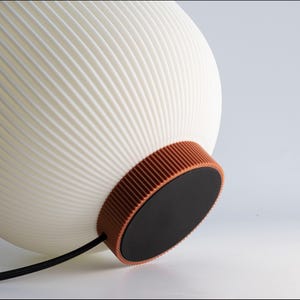May include: A white, ribbed, spherical lamp with a brown band and a black base. The lamp has a modern design and a black power cord. The lamp is a decorative lighting fixture.