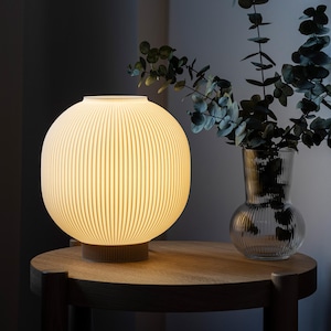 May include: A glowing, spherical table lamp with vertical ribbed detailing. The lamp sits on a dark wooden base and is placed on a round wooden table. A clear glass vase with green foliage is next to the lamp.