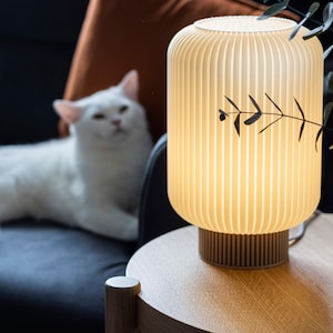 Puede incluir: Una lámpara de mesa crema, acanalada, con una pantalla cilíndrica y una base de madera. La lámpara está iluminada, emitiendo un brillo cálido. Una ramita de hojas está en la pantalla. Un gato blanco está en el fondo.