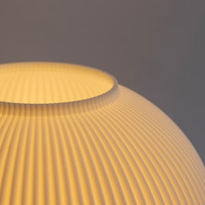 May include: Close-up of a white, ribbed lampshade with a rounded shape. The shade has a textured surface with vertical lines, and the interior glows with a warm light. The design is modern and minimalist.