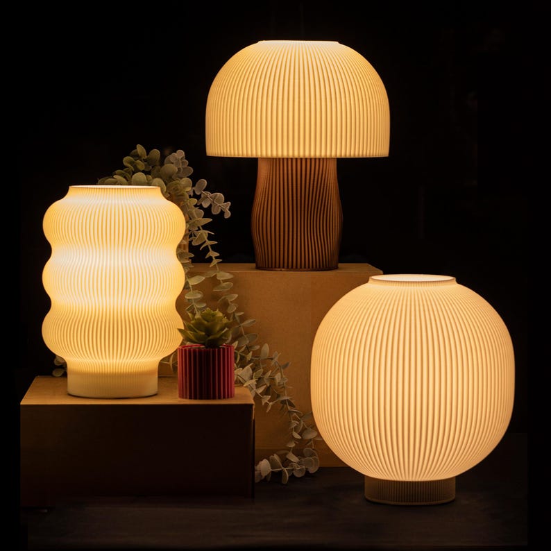May include: Three illuminated lamps with ribbed shades and warm light. The lamps have different shapes: a mushroom-shaped lamp, a wavy lamp, and a spherical lamp. A small red pot with a succulent plant and greenery are also present.
