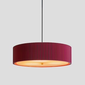 May include: A cylindrical pendant light fitting featuring a deep magenta, vertically ribbed exterior shade. The bottom of the drum-shaped lamp is covered by a circular diffuser that emits a warm, soft glow, suspended by a single thin black cord.