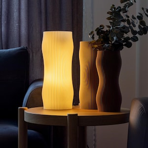 May include: A cream-coloured lamp with a ribbed design is illuminated on a wooden side table. Two brown vases with vertical grooves are also on the table. The lamp and vases are decorative home accents, creating a warm atmosphere.
