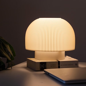 May include: A cream-colored table lamp with a ribbed, dome-shaped shade, emitting a warm glow. The lamp sits on a white base, resting on two books. A laptop is partially visible in the foreground. The lamp's design is modern and minimalist.