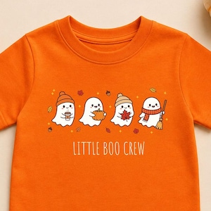 Könnte beinhalten: Ein orangefarbenes T-Shirt mit kurzen Ärmeln und einer Grafik von vier weißen Geistern. Die Geister tragen herbstliche Accessoires und halten saisonale Gegenstände. Unter den Geistern steht der Text LITTLE BOO CREW in weißer Schrift.