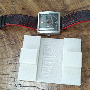 May include: A square-faced chronograph wristwatch with a dark grey dial, silver-tone hands, and a black perforated leather strap with red lining. Below the watch sits a white rectangular display stand embossed with the word HEUER.
