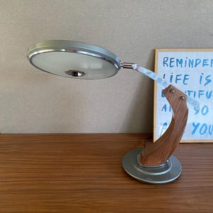 May include: A desk lamp featuring a circular, frosted white light head with a chrome rim, supported by a curved wooden arm and a circular silver-toned base. The lamp has a metallic finish and a distinct, arched structure connecting the base to the head.