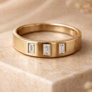 May include: A gold band ring with three rectangular, clear gemstones set across the top. The ring has a flat, wide band and a polished finish. The ring is displayed on a neutral-toned surface.