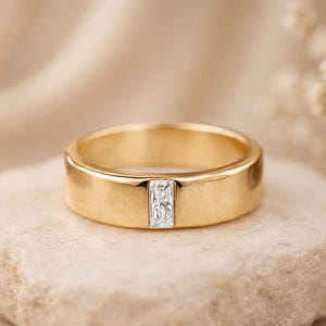 May include: A gold-colored ring with a rectangular, clear stone set in the center. The ring is displayed on a light-colored, textured stone, with a soft, neutral background.