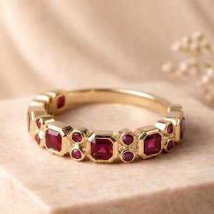 Può includere: Un anello d'oro con pietre preziose rosse quadrate e rotonde alternate. L'anello è posto su una superficie dai toni neutri, con uno sfondo morbido e sfocato. Il design dell'anello è simmetrico.
