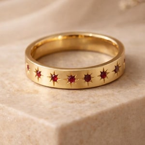 May include: A gold-colored ring with a series of star-shaped cutouts, each containing a small, red gemstone. The ring is set on a neutral-toned surface, showcasing its design. The ring's band is approximately 0.25 inches wide.