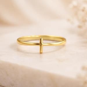 May include: A gold-colored ring with a cross design. The ring is set against a neutral background, highlighting the simple yet elegant design. The cross is centered on the band, creating a minimalist aesthetic. The ring is likely made of gold or a gold-toned metal.