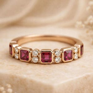 May include: A rose gold band ring featuring a repeating pattern of three square-cut deep pink rubies alternating with pairs of small round white diamonds. The gemstones are set in a bezel style along the top half of the polished metal ring band.