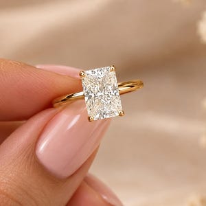 Op de afbeelding: Een gouden ring met een grote, rechthoekige, heldere edelsteen in radiant-slijpvorm, vastgehouden door een zetting met vier pootjes. De ringband is dun, gepolijst en rond, en toont een klassiek ontwerp met een opvallende, gefacetteerde steen.