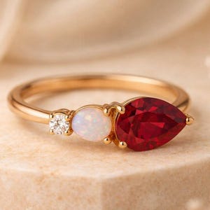 以下が含まれることがあります： 洋梨型の深い赤色の宝石、楕円形の白い宝石、小さな丸い透明な宝石をあしらったゴールドリング。 リングはニュートラルな表面に置かれ、ジュエリーのデザインと色を際立たせています。