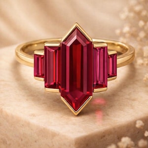 May include: A gold ring featuring a central elongated hexagonal red gemstone flanked by two smaller rectangular red gemstones on each side, all set in a polished gold band with a clean, symmetrical arrangement of the five total stones.