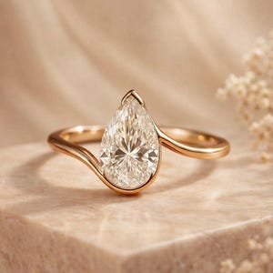 Könnte beinhalten: Ein Ring aus Roségold mit einem birnenförmigen Mittelstein in einer Zargenfassung. Die Ringschiene hat ein glattes, geschwungenes Design, das um die Basis des Steins verläuft und eine durchgehende, fließende Silhouette mit polierter Oberfläche bildet.