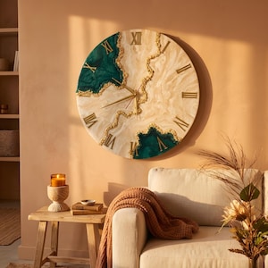 Puede incluir: Un gran reloj de pared redondo con un diseño crema y verde esmeralda. El reloj presenta números romanos dorados y detalles dorados. El reloj está colgado en una pared de color melocotón. El diámetro del reloj es de unos 61 cm.